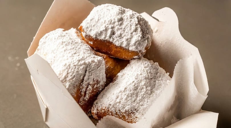 A white paper box filled with several golden-brown beignets, heavily dusted with powdered sugar.
